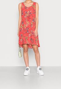 Robe trapèze fleurie avec un fond corail, ornée de motifs verts et bleus, col rond et sans manches. Associée à des baskets blanches.