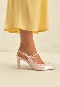 Scarpa con tacco alto rosa, con cinturino a slingback e dettagli in punta argentata a onda. Presenta un fiocco e un design con tacco snello.