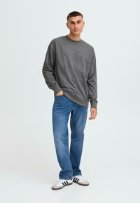 Ung mann står avslappet iført en grå genser, blå dongeribukser og hvite joggesko med svarte striper mot en ensfarget bakgrunn.