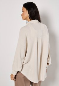 Beige, long-sleeved top in a textured waffle knit, featuring a collared neckline and a relaxed fit with a curved hem.