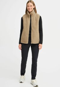 Gilet en polaire beige avec col haut et fermeture éclair à l'avant ; porté sur un pull noir et associé à un pantalon sombre et des baskets blanches.