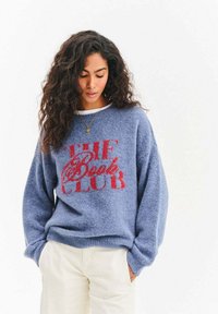 Niebieski oversize'owy sweter wykonany z miękkiego, puszystego materiału, z napisem "THE Book CLUB" na czerwono. Ściągacze na mankietach i dole, luźny krój.