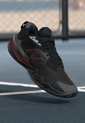 Chaussure de sport noire avec une tige texturée, des accents rouges et une semelle en caoutchouc. Présente un profil bas et un embout renforcé.