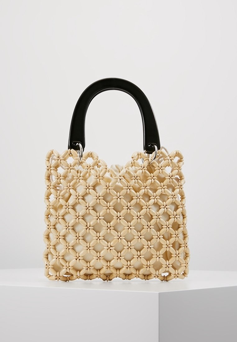 Sac à main beige en tissu tissé avec un motif floral, présentant une finition texturée et une poignée noire brillante. Forme compacte et structurée.