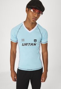 Ανοιχτό μπλε αθλητικό πουκάμισο με V λαιμόκοψη, λευκές λεπτομέρειες, μαύρο λογότυπο "URTRN" και τον αριθμό "32". Μοντέλο που φοράει διάφανα γυαλιά ηλίου και μαύρο παντελόνι.