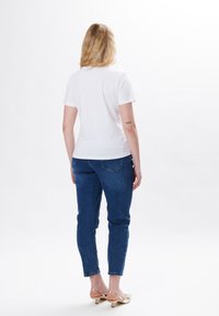 Hvid kortærmet t-shirt, blå denim jeans og guld hæle sandaler. Bagfra ses et enkelt design uden synlige mønstre eller logoer.