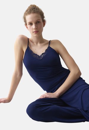 Jeune femme aux cheveux bouclés portant un caraco bleu marine orné de dentelle et un pantalon, assise avec un bras reposant sur le sol.