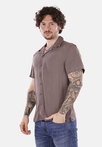Camicia a maniche corte con bottoni in tessuto marrone chiaro dal tocco morbido. Presenta un colletto da campeggio e dettagli minimi. Indossata con jeans blu.