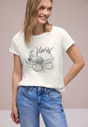 Smilende kvinde med blondt hår iført en hvid T-shirt med en sort stregtegnet blomst og teksten "joy floral", kombineret med blå jeans.