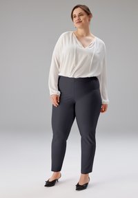 Weiße Bluse mit V-Ausschnitt und verstellbaren Kordelzügen, kombiniert mit taillierten dunklen Hosen. Elegante schwarze spitze Schuhe vervollständigen das Outfit.