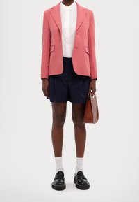 Blazer rosa sartoriale, camicia bianca con colletto, pantaloni corti blu navy e scarpe nere con suole spesse. Borsa marrone e calzini bianchi visibili.