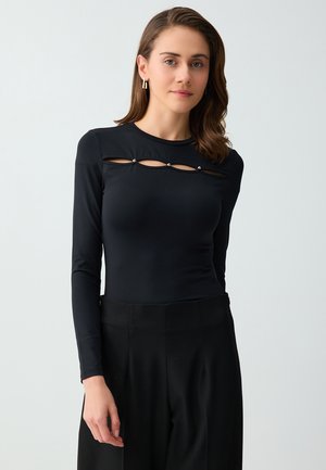 Vrouw met bruin haar tot op de schouders, gouden oorbellen, een zwarte top met lange mouwen met uitsnijdingen en parelknopen op de borst, en een zwarte broek.