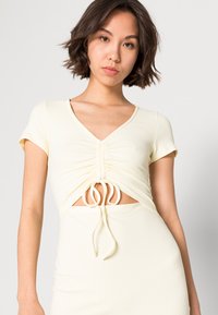 Top cropped amarelo com nervuras, decote em V, mangas curtas e um detalhe central de cordão. O design apresenta um recorte na cintura.
