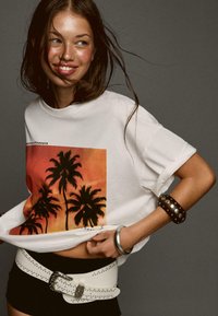 White t-shirt featuring a sunset graphic with palm trees, paired with black shorts and a studded white belt. Accessories include a silver bracelet.