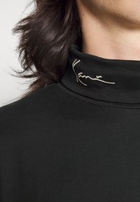 Schwarzes Rollkragenshirt aus glattem Stoff. Mit einem gestickten Signaturdetail in Weiß am Kragen. Nahaufnahme.