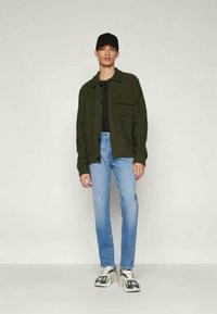 Olivgrön knappjacka med bröstficka, kombinerad med ljusblå jeans och vita sneakers med mörka detaljer. Svart keps bärs.