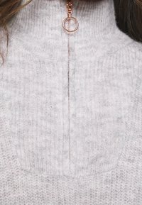 Lichtgrijze zip-up sweater met hoge kraag, voorzien van een geribbelde textuur en een roségouden ritssluiting. Duidelijke naaddetails langs de voorkant.