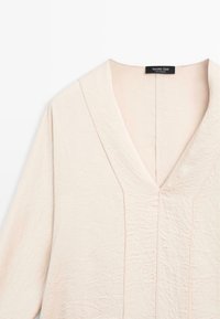 Hellrosa Bluse aus strukturiertem Stoff, mit tiefem V-Ausschnitt und langen Ärmeln mit dezenten Nahtdetails. Etikett am Hals sichtbar.
