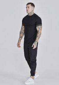 Sort tætsiddende t-shirt med korte ærmer, med gyldne accentstriber. Iført med sorte joggers og hvide sneakers. Tatoveringer synlige på armene.