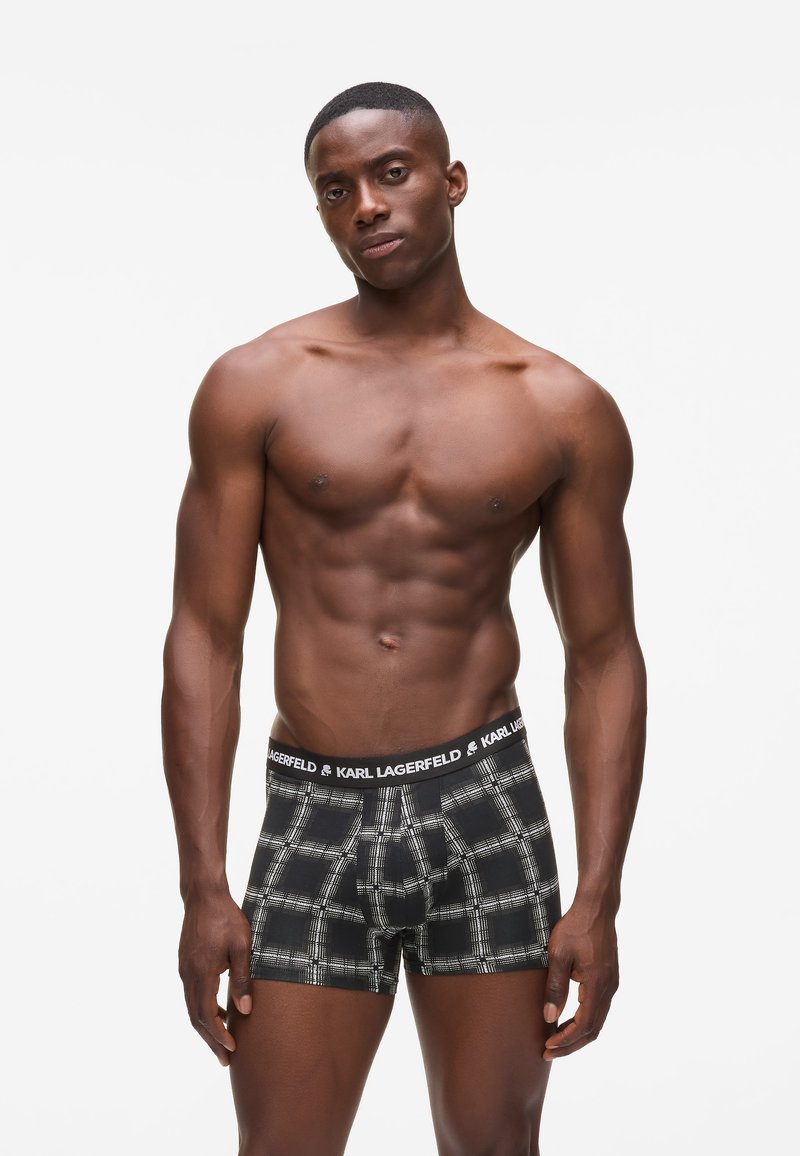Izmos férfi rövid hajjal, fekete-fehér kockás boxeralsót visel, amelynek derékrészén Karl Lagerfeld logó látható, fehér háttérrel.
