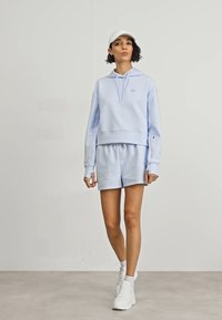 Hanorac scurt bleu deschis cu șnururi, combinat cu pantaloni scurți asortați. Are un material moale și un design minimal. Tenisi albi și o șapcă.