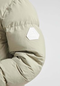 Lys beige puffy jakkeærme med quiltet tekstur og et karakteristisk hvidt sekskantet logo-patch på albueområdet.