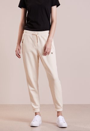 Jogginghosen in Cremefarben mit metallischen Reißverschlusstaschen, elastischem Bund und Bündchen. Kombiniert mit einem schwarzen T-Shirt und weißen Turnschuhen.