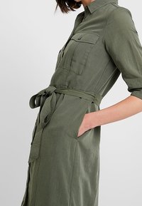 Olivově zelené šaty košilového střihu s krátkým zapínáním, vyrobené z lehkého materiálu, s páskem v pase, dvěma náprsními kapsami a dlouhými rukávy.