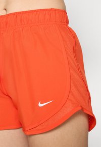 Orange athletic shorts with a stretchy waistband, side mesh panels, and a white Nike logo on the lower left. Lightweight fabric texture.