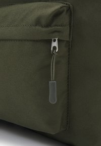 Bolsa de tela color verde olivo con una superficie texturizada, que presenta un cierre plateado y una pestaña de tira gris. El diseño incluye un bolsillo frontal.
