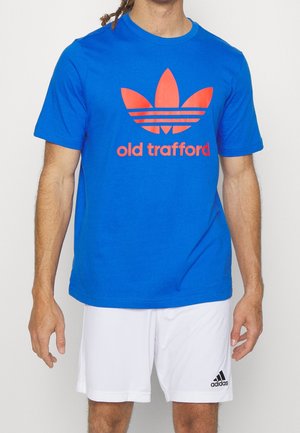 Mann trägt ein blaues T-Shirt mit rotem Adidas-Logo und dem Schriftzug "Old Trafford", kombiniert mit weißen Adidas-Shorts.