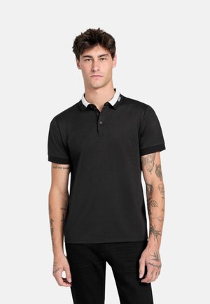 Giovane con capelli castani che indossa una polo nera con colletto bianco, in piedi davanti a uno sfondo chiaro e semplice.