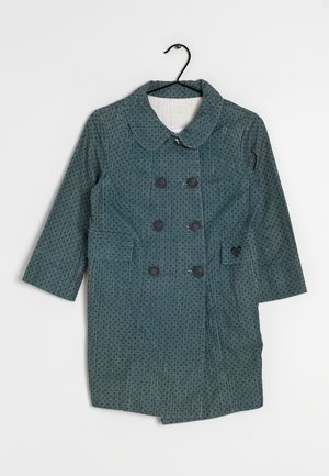 Doppelreihiger tealfarbener Mantel mit schwarzen Punkten, mit Kragen, zwei Fronttaschen und vier schwarzen Knöpfen. Hängt an einem schwarzen Haken.
