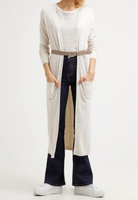 Lång krämfärgad cardigan med fickor, bärs över en randig topp, åtdragen i midjan med ett brunt bälte, ihop med mörkblå utsvängda jeans och vita sneakers.