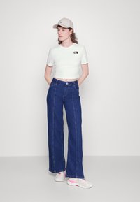 Camiseta blanca ajustada con logotipo, jeans de pierna ancha azul y gorra color crema. Calzado con acentos en blanco y rosa. Texturas de tela suaves.