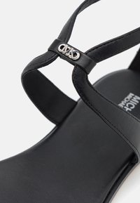 Bride de sandale en cuir noir avec détail du logo « MK » argenté sur la bride en T au-dessus d’une semelle noire avec le nom de la marque embossé.