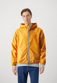 Gul zip-up jakke med hette, elastiske mansjetter og grå midjebånd. Har en logo på venstre bryst og en glatt, vannavstøtende materiale.