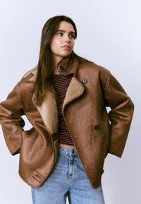 Mujer joven vestida con una chaqueta de borreguillo marrón oversized, camiseta de rayas y jeans de un azul claro, de pie con las manos en las caderas contra un fondo blanco.