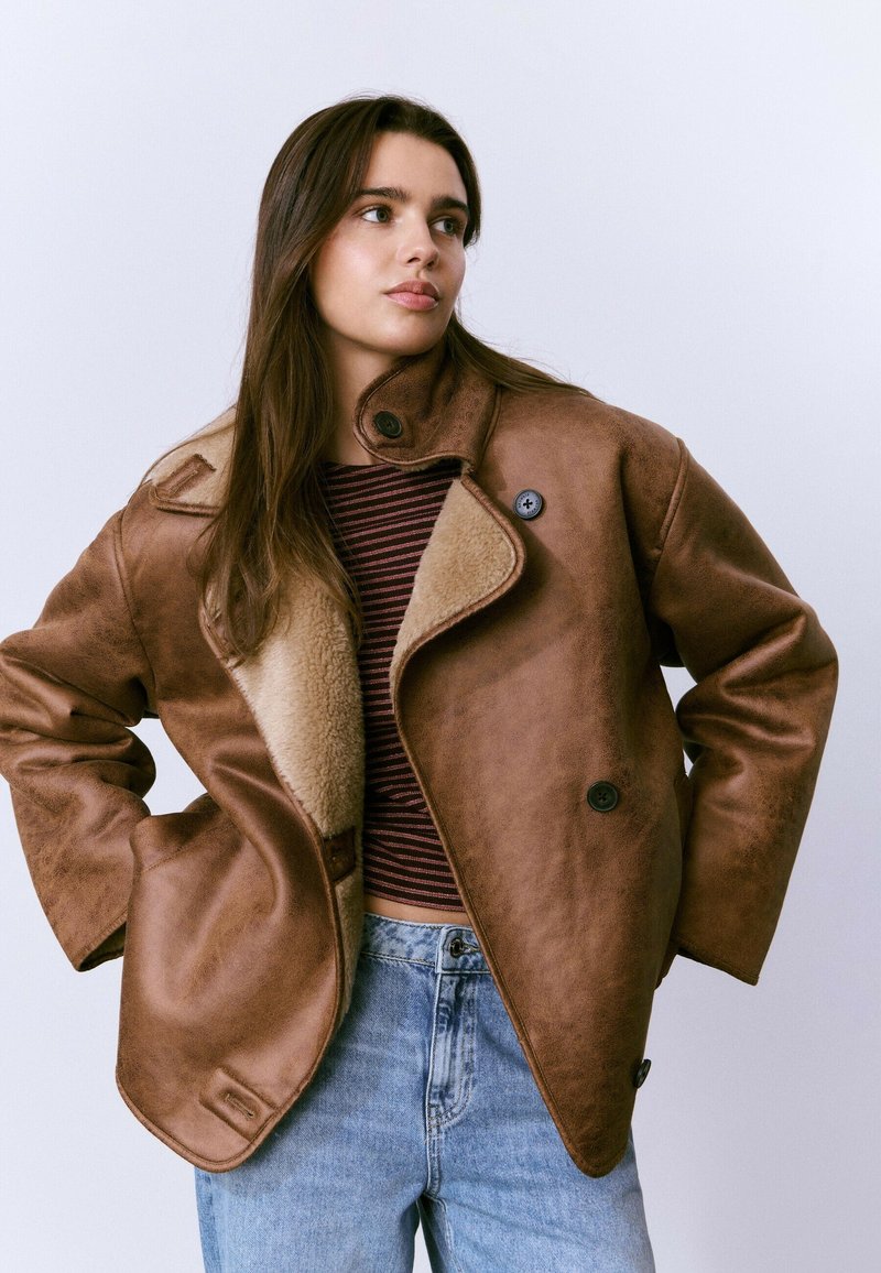 Mujer joven vestida con una chaqueta de borreguillo marrón oversized, camiseta de rayas y jeans de un azul claro, de pie con las manos en las caderas contra un fondo blanco.