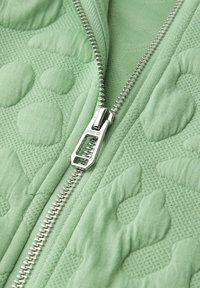 Lähikuva hopeisesta vetoketjusta vaaleanvihreällä tikatulla kankaalla, jossa on kohotetut, tekstuurilliset kukkakuviot.