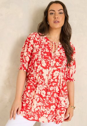 Kobieta ubrana w luźną czerwoną bluzkę z kremowym kwiatowym wzorem, plisowanymi detalami i bufkami o długości do łokcia, w połączeniu z białymi spodniami.