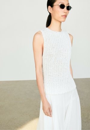 Femme portant des lunettes de soleil noires et un top en maille blanc sans manches avec un pantalon blanc ample, debout dans un espace intérieur bien éclairé et minimaliste.