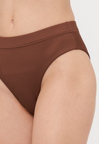 Rjavi rebrasti bikini spodnji del z širokim pasom, visokimi izrezi na nogah in gladko teksturo. Ima prilegajočo se obliko in minimalne šive.