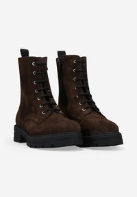 Botas de tobillo de ante marrón con cordones, suelas de goma negra gruesa y cremalleras laterales, colocadas una al lado de la otra sobre un fondo blanco.