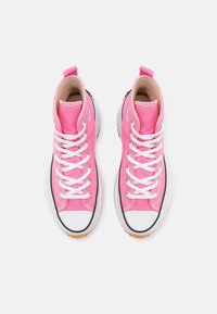 Vaaleanpunaiset korkeat canvas-tennarit, joissa on valkoiset nauhat ja kumiset varvaskappaleet, nähtynä ylhäältä valkoista taustaa vasten.