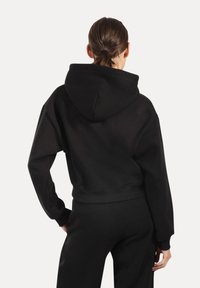 Haut noir court avec capuche ; présente une coupe ample et des poignets côtelés. Tissu doux, design minimal, montré de dos.