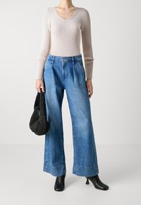 Femme portant un haut à manches longues côtelé rose clair, un jean bleu taille haute à jambes larges, des bottines noires à talons, tenant un sac à main noir tissé.