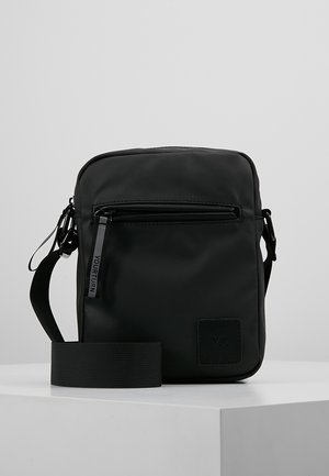 Sac bandoulière noir en nylon durable. Il comprend une poche avant zippée et une sangle en tissu réglable. Texture mate lisse avec un écusson logo.