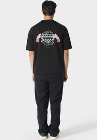 Sort bomulds t-shirt med ternede flag og et blomstermotiv på ryggen, med teksten "PAS DE MONACO" i fed skrift.