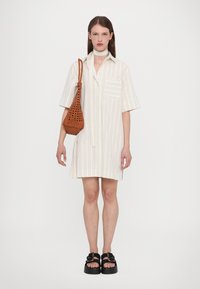 Abito a maniche corte con colletto, caratterizzato da righe verticali in crema e beige, con taschino sul petto. Abbinato a una borsa intrecciata marrone e sandali con piattaforma.
